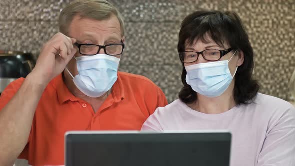 Couple Senior in Protective Masks Using Laptop at Home for Online Shopping alt