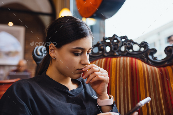 Pretty young girl resting on a big soft chair in a cafe, chatting on the phone