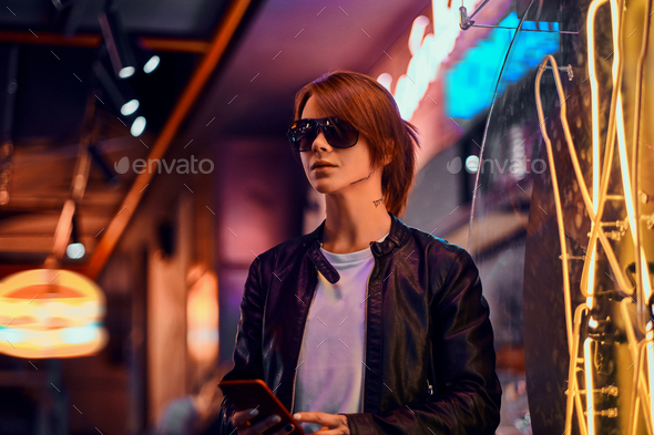 Stylish redhead girl holding a phone while standing in the night on the street. - Stock Photo - Images