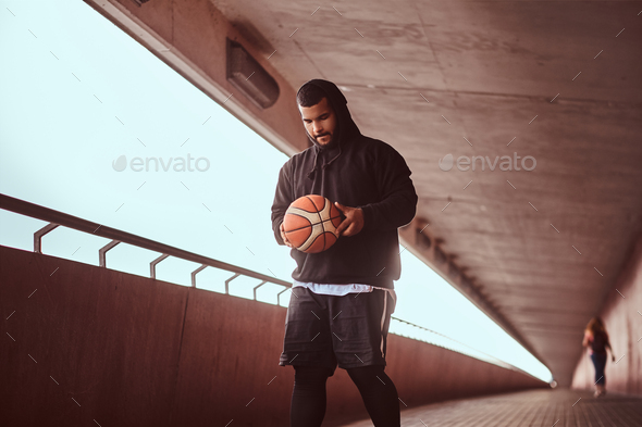Dark-skinned guy holds a basketball while standing on a footway under bridge. - Stock Photo - Images