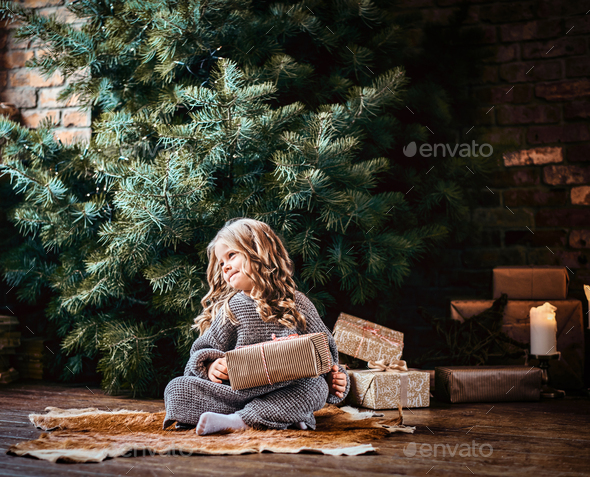 Cute little girl with blonde curly hair sitting on a floor next to the christmas tree - Stock Photo - Images