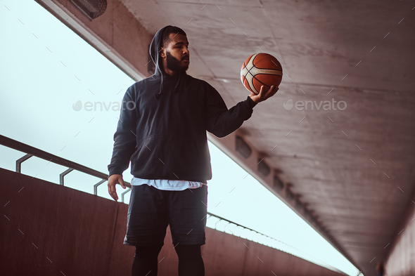 Dark-skinned guy holds a basketball while standing on a footway under bridge.