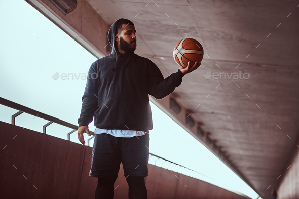 Dark-skinned guy holds a basketball while standing on a footway under bridge. - Stock Photo - Images
