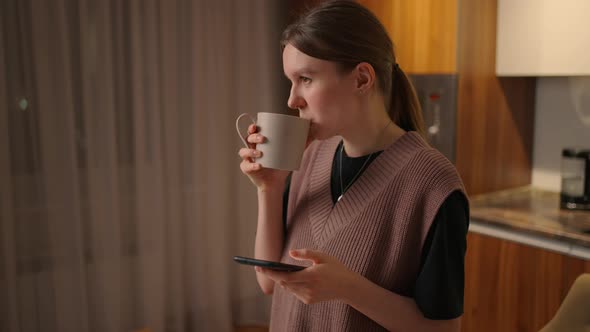 A Young Beautiful Woman at Home in the Kitchen Looks at Her Cell Phone and Drinks Coffee alt