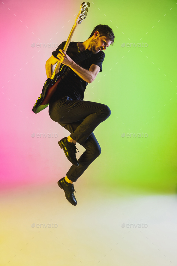 Young caucasian musician playing bass guitar in neon light on pink-green background