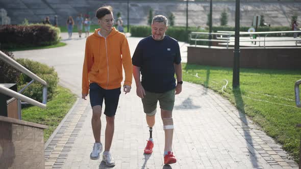 Wide Shot Portrait Positive Teenage Son Walking with Mature Father Amputee in Urban City Talking alt