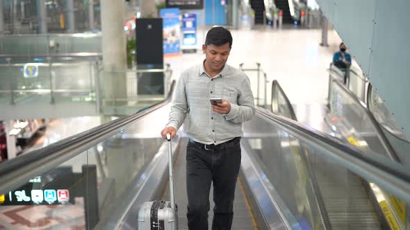 Young man with suitcase standing on the moving walkway while using smartphone.