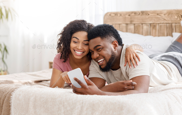 Happy black couple using smartphone on bed - Stock Photo - Images