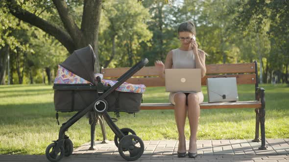 Wide Shot of Concentrated Young Businesswoman Sitting in Summer Park with Baby Stroller, Laptop, and alt