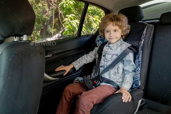 Happy cute boy of elementary age in denim jacket and brown pants sitting in car - Stock Photo - Images