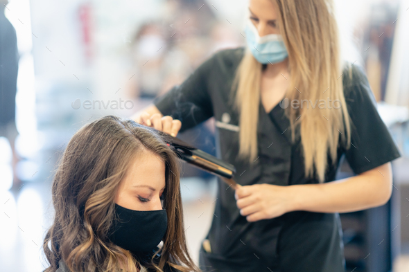 Hairdresser, protected by a mask, combing her client's hair with a hair iron in a salon - Stock Photo - Images