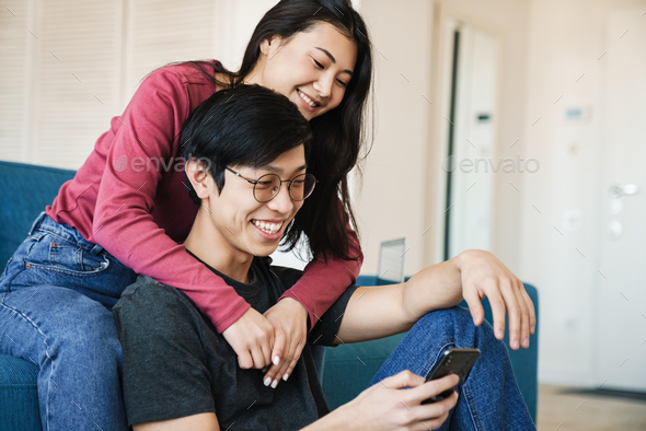 Photo of asian couple using mobile phone while sitting at cozy room
