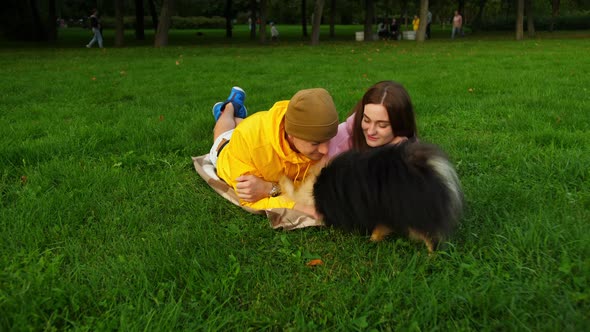 Young Guy and Girl Play with a Little Cute Dog Lying on the Grass in the Park alt