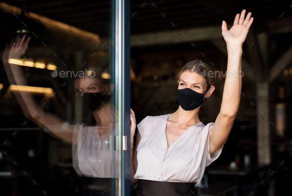 Portrait of waitress with face mask standing at the door in restaurant, waving
