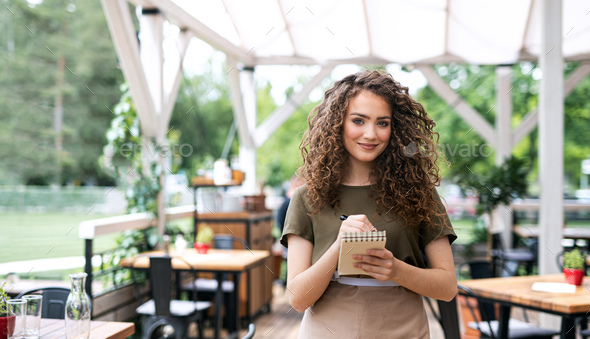 Waitress with order pad standing outdoors on terrace restaurant, looking at camera