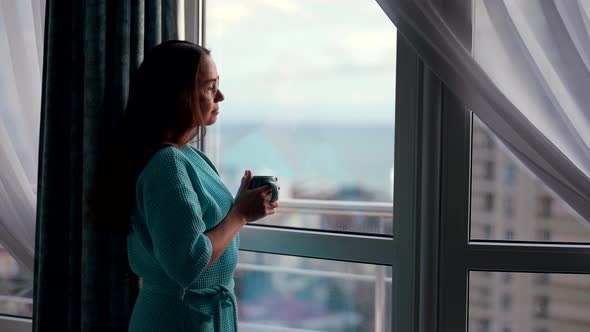 an Attractive Woman at Home in the Morning in a Bathrobe Stands at the Window and Drinks a Hot Drink alt