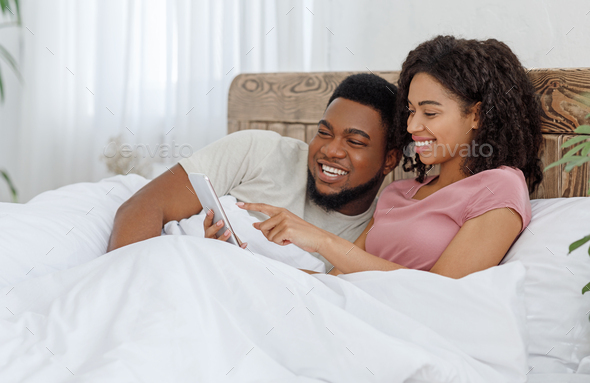 Happy black couple watching photos on phone in bed