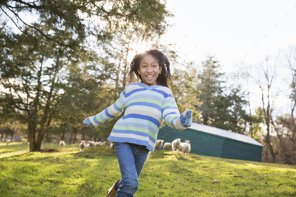 A young girl in a blue stripey top running in a field of sheep at an animal sanctuary.