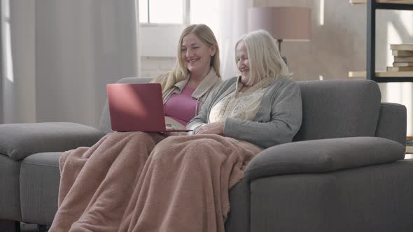 Wide Shot Portrait of Two Laughing Senior Caucasian Women Surfing Social Media on Laptop Talking alt