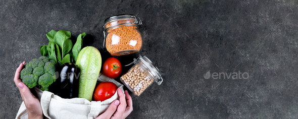 Zero waste concept. Female hands holding vegan vegetables in cotoon reusable bag, glass jars with chickpea, lentils on dark slate table. Top view, copy space, flat lay. Banner image for website