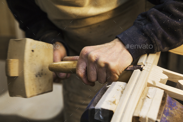 Close up of a man standing at a work bench in a carpentry workshop, working on a wooden chair with a wooden mallet and chisel.