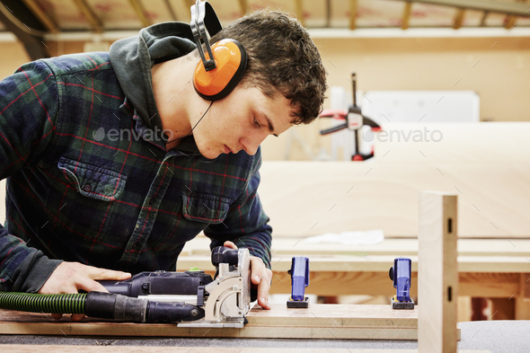 A young man using tools to shape a piece of wood in a furniture workshop. - Stock Photo - Images