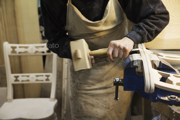 Man standing at a work bench in a carpentry workshop, working on a wooden chair with a wooden mallet and chisel.