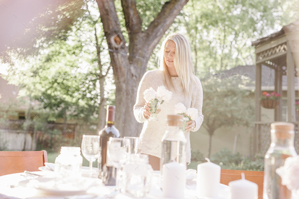 Blond woman setting a table in a garden, candles and vases with pink roses.