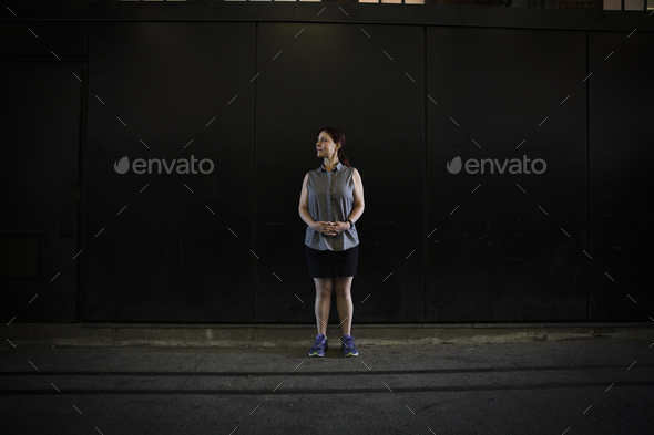 A woman in a sleeveless shirt and shorts with running shoes, standing in shadow outside a building on a street.