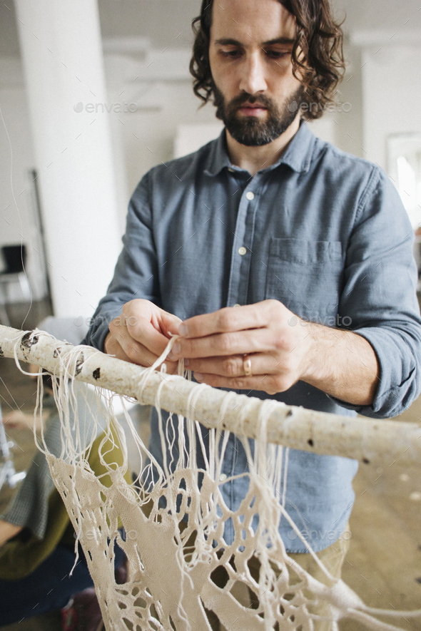 An artist working on an art piece hanging on a frame, knotting and weaving threads.
