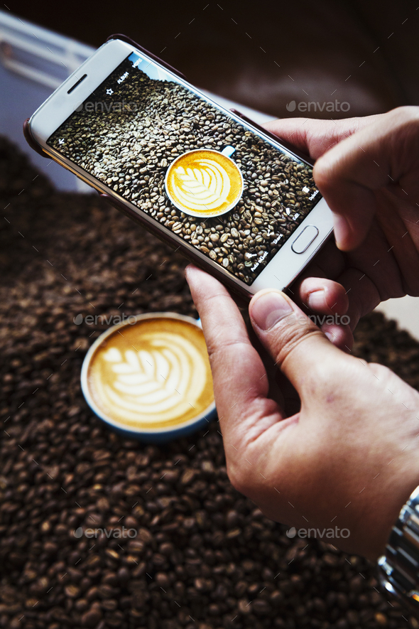 Specialist coffee shop. A person taking a picture with a smart phone of a cup of coffee on top of a heap of roasted coffee beans.