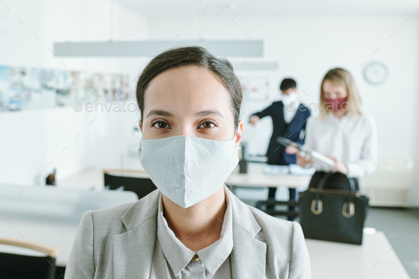 Young mixed-race businesswoman in protective mask looking at you in office