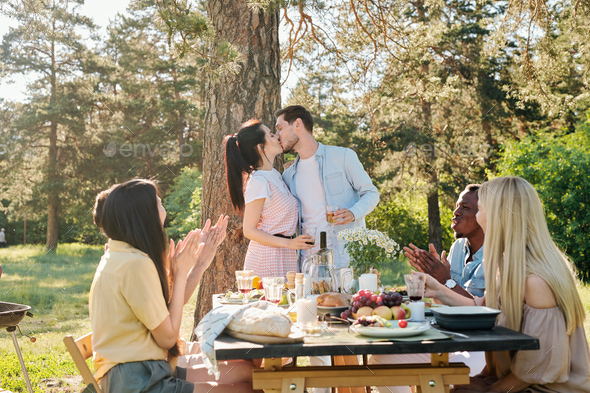 Amorous couple kissing under pine tree while their friends congratulating them