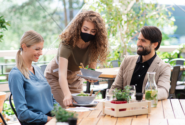 Waitress with face mask serving happy couple outdoors on terrace restaurant