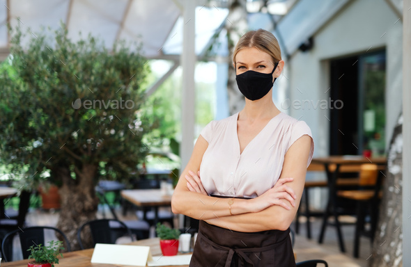 Waitress with face mask standing outdoors on terrace restaurant, arms crossed
