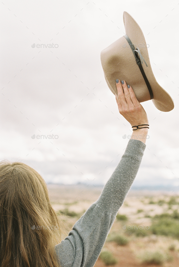 Rear view of a woman standing in the desert, holding a hat, her arms raised up in the air.