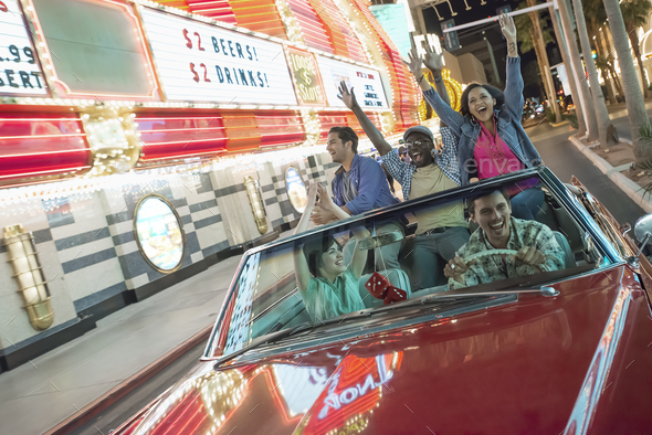 Friends in a red convertible celebrating with waving arms, driving in a city lit with neon signs.