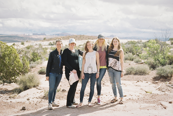 Group of five women, friends standing side by side in a desert landscape smiling.