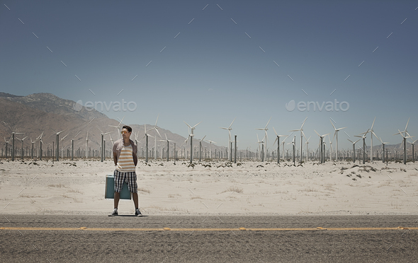 A man standing with a suitcase on the side of a highway, with wind turbines in the background.