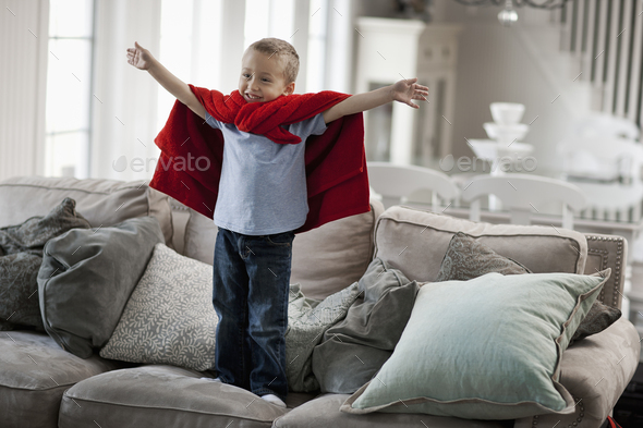 A child standing wearing a red cape, arms raised in a a superhero pose.