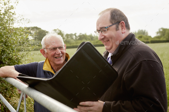 Two men standing looking over a farm gate, one with an open file and paperwork.