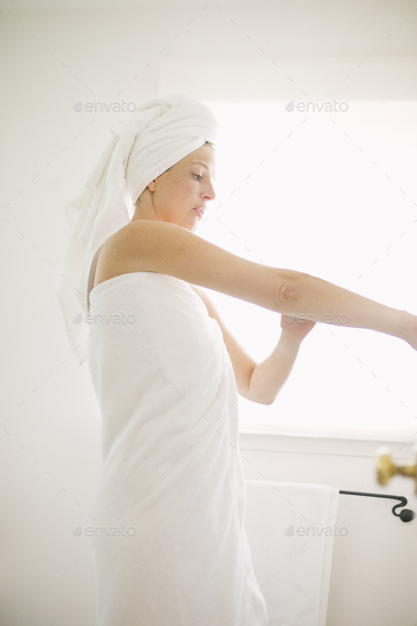 Woman wrapped in a white towel standing in a bathroom, applying lotion to her arm.