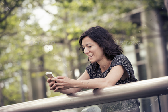 A woman leaning on a rail in a city park checking her cell phone - Stock Photo - Images