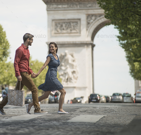 A couple hand in hand carrying shopping bags and crossing the road by an arch monument. - Stock Photo - Images