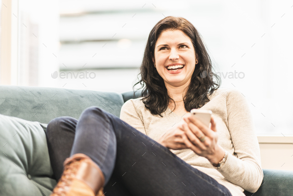 A woman seated with her feet up on a sofa, looking at her smart phone.