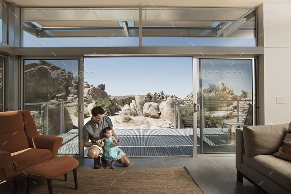 A man and a young child in an ecohouse, a home with large glass walls and view out over the rocky landscape.
