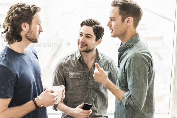 Three men standing talking, one with a cup of coffee, one with a smart phone.