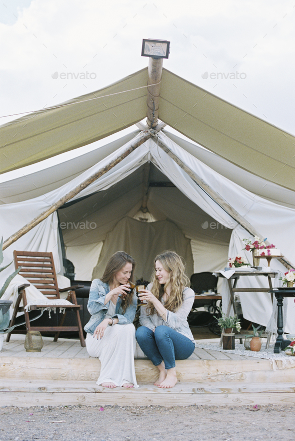 Two smiling women sitting outside a large tent laughing and having a glass of wine.