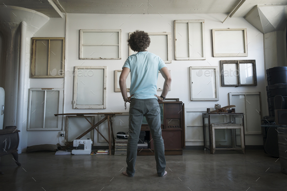 Loft decor. A wall hung with pictures in frames, reversed to show the backs. A man with hands on his hips looking at the wall.