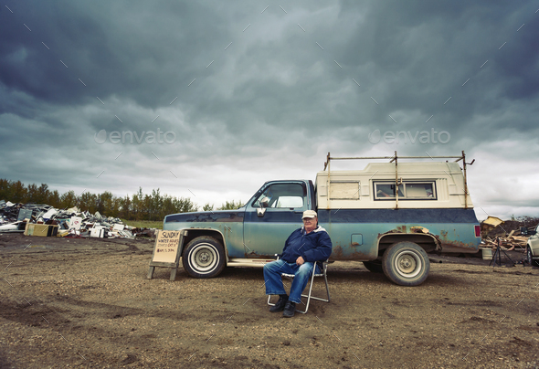 A mature man seated in a chair by his pick up truck. Piles of waste, scrap metal and wood objects.
